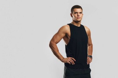 Siyah spor kıyafeti giyen genç sporcu elleri kalçasında tarafsız gri bir zemine karşı güven içinde poz veriyor. Form, güven, kararlılık ve sağlık konuları için mükemmel..