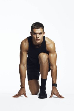 Kararlı genç bir erkek, bir stüdyoda sürat koşusu için hazırlanıyor, güçlü ve odaklı, beyaz arka planda izole. Fitness pazarlama ve motivasyon içeriği için ideal.