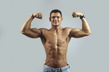 Atletik genç adam kaslarını güvenli bir şekilde bir stüdyo ortamında esnetiyor, güç ve odaklanma gösteriyor, gri arka planda izole edilmiş. Fitness markalaşma ve reklam kampanyaları için ideal..