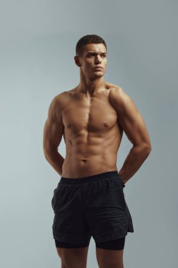Kendine güvenen kaslı, formasız adamın dikey fotoğrafı. Gri arka planda poz veren, elleri arkada olan, başka yöne bakan bir adam.