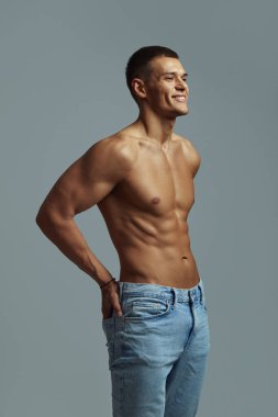 Çekici genç bir adam formda ve atletik bir fiziğe sahip, mutlu duruyor ve basit gri bir arka plana karşı pozitifliğini ve sağlığını ifade ediyor. Fitness ve yaşam tarzı temaları için mükemmel.