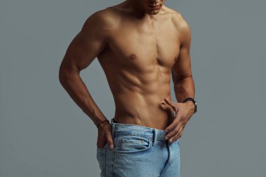 Atletik tişörtsüz bir erkek, gri arka planda mavi kot pantolon giyerken vücut ve kas yapısını ön plana çıkarır..