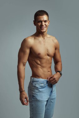 Sıradan bir kotla poz verirken kasları ve kendine güveni olan, cana yakın, üstsüz bir adam. Olumlu ifadesi, nötr zemin karşısında izole edilmiş sağlık, sağlık ve yaşam tarzı ideallerini aktarıyor.