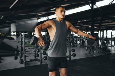 Spor giyim bölümündeki yakışıklı vücut geliştirme uzmanı modern spor salonundaki kasları dambıllarla temizliyor. Vücut geliştirme, fitness ve sağlık hizmeti kavramı.