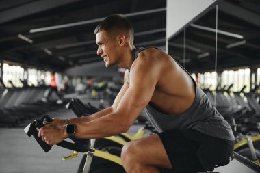 Modern jimnastik salonunda sabit bisiklet üzerinde egzersiz yapan yakışıklı adamın yan görüntüsü. Kolunu sıkıca tutup ileriye bakıyor. Spor ve sağlık hizmetleri kavramı.