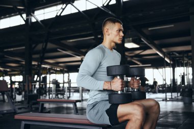 Geniş bir spor salonundaki bir bankta oturan halterli bir sporcunun yatay fotoğrafı. Yoğun bir üst vücut çalışmasından önce bir hazırlık anında güç ve disiplin yakalanır.