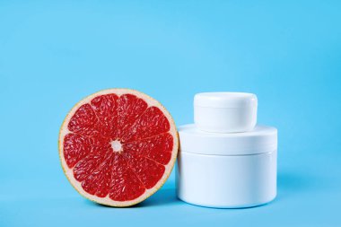 Taze bir greyfurt dilimi beyaz tenli krema kavanozlarının yanında parlak mavi bir zemin üzerinde oturur, doğal güzelliği ve kişisel bakım kavramlarını ön plana çıkarır..