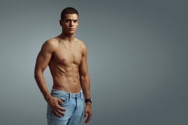 Atletik genç adam modern bir stüdyoda üstsüz poz veriyor. Sağ tarafta boş alan olan gri arkaplana karşı kaslı fiziğin ve gelişigüzel cazibenin vurgulanması