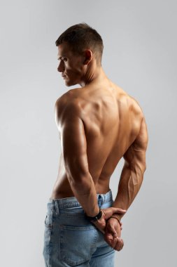Uyumlu erkek, kaslarını ve sırt ve kol kaslarını sergilerken gri arka planda izole bir şekilde duruyor. Gücü ve tarzı somutlaştırıyor. Fitness ve moda pazarlaması için ideal..