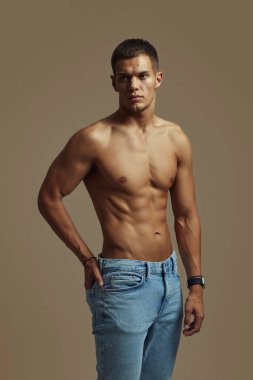 Kaslı genç adam kendinden emin bir şekilde üstsüz poz veriyor, formda olduğunu ve günlük kot pantolon giydiğini gösteriyor. Fitness, moda, güven, erkeksilik, güç ve kişisel tarz gibi konular için ideal.