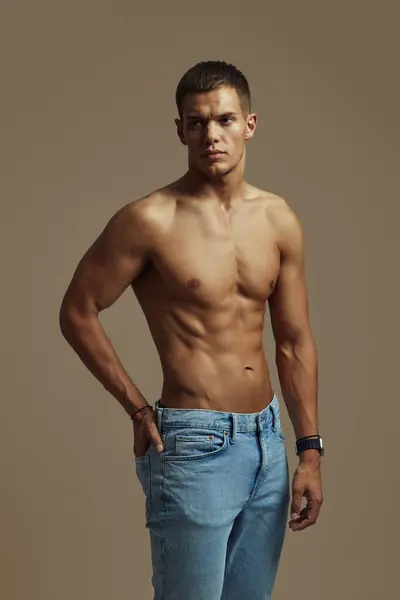 Kaslı genç adam kendinden emin bir şekilde üstsüz poz veriyor, formda olduğunu ve günlük kot pantolon giydiğini gösteriyor. Fitness, moda, güven, erkeksilik, güç ve kişisel tarz gibi konular için ideal.