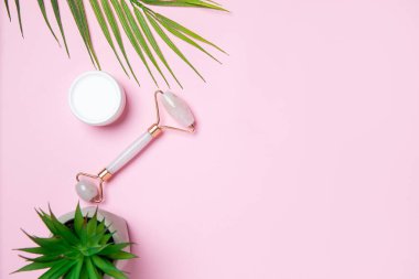 Yeşil palmiye yaprakları ve taze ve rahatlatıcı bir bitki içeren pembe arka planda yüz silindiri ve krem de dahil olmak üzere cilt bakımının en önemli unsurları..