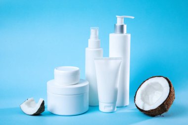 Parlak beyaz kaplarda doğal cilt bakımı ürünlerinin toplanması. Temiz mavi arkaplan üzerine kurulmuş taze hindistan cevizi yarıları, organik güzelliği ve kendine bakımı vurguluyor..