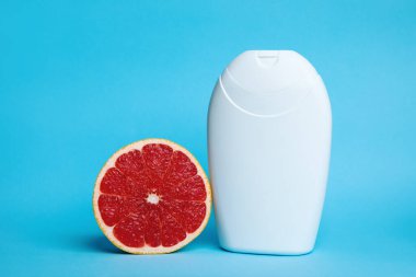 Taze bir greyfurt yarısı ve parlak beyaz bir şişe canlı mavi bir zemin üzerine kurulur, tazelik ve temizliği çağrıştırır. Cilt bakımı, sağlık ve doğal güzellik kavramları için ideal..