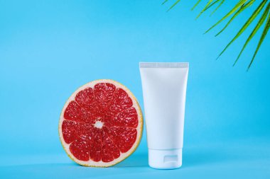 Canlı mavi bir arka planda, boş bir deri tüpünün yanında taze bir greyfurdun parlak görüntüsü. Doğal güzellik, sağlık ve sağlık ürünlerini canlandırıcı bir bağlamda desteklemek için idealdir..