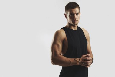 Siyah kolsuz tişörtlü kararlı genç bir adam beyaz arka plana karşı fiziğini sergiliyor, gücü ve güveni simgeliyor. Vücut geliştirme, fitness ve sağlık hizmeti kavramı.