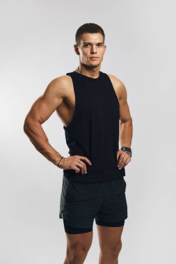 Kaslı genç bir adam, spor kıyafeti içinde, güçlü ve kararlı bir şekilde poz veriyor, profesyonel bir stüdyoda, temiz gri bir arkaplanda çekiliyor..