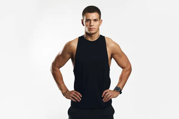 Siyah kolsuz bluz giyen kaslı genç adam, beyaz arka plana karşı kararlı ve güçlü görünerek kalça üzerinde el ele tutuşurken güven içinde poz veriyor. Vücut geliştirme, fitness ve sağlık hizmeti kavramı.