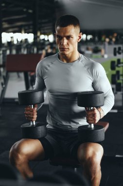 Modern spor salonunda iki ağır dambılla antrenman yapmaya hazırlanan kararlı bir adamın dikey fotoğrafı..