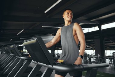 Spor kıyafetli yakışıklı beyaz adam koşu bandında yürürken gülümsüyor, iyi donanımlı spor salonunda kardiyo seansına hazırlanıyor. Sağlıklı bir adam, sağlık ve zindelik üzerine çalışırken, gözünü kaçırmadan yürür..