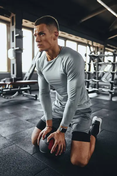 Güçlü, tanımlı bir vücudu olan yakışıklı bir adamın dikey fotoğrafı tıp topu eğitiminden sonra dinlenmek için biraz zaman alır. Modern aktif giysiler giyerek spor salonunda diz çöküp ciddi bir ifade takınıyor..