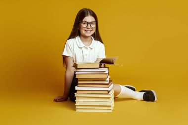 Gözlük takan ve okul üniforması giyen gülümseyen çocuk, öğrenmeyi sevdiğini ifade eden bir yığın kitabın yanında güven içinde oturuyor. Eğitim terfileri ya da okula dönüş kampanyaları için ideal..