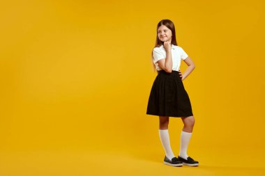 Klasik siyah beyaz okul üniformalı neşeli bir genç kızın tüm vücudunun yatay fotoğrafı canlı sarı bir arkaplan karşısında duruyor ve boş alana bakıyor. Eğitici içerik için mükemmel