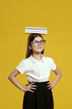 Mutlu çocuk kafasında bir yığın kitabı dengelerken yukarı bakıyor, oyuncu bir sahnede odaklanma ve kararlılık gösteriyor. Eğitimsel içerik ve okulla ilgili reklamlar için ideal.