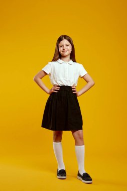 Neşeli bir çocuk, parlak sarı bir arka planda, klasik okul üniforması içinde, elleri kalçalarının üzerinde güvenle ayakta duruyor. Eğitim terfileri ve okul kampanyaları için ideal.