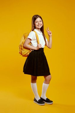 Okul üniformalı neşeli bir gencin dikey fotoğrafı canlı bir sırt çantası taşıyor, sarı zemine karşı barış jesti yaparken sevinçle duruyor. Okul kampanyaları için ideal..
