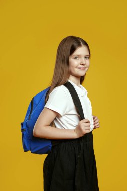 Mavi bir sırt çantası tutan neşeli bir genç, parlak sarı bir arka planın önünde güvenle duruyor. Eğitici reklamlar ve okula dönüş promosyonları için mükemmel..