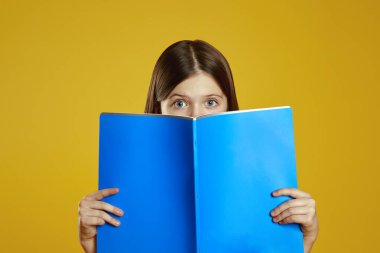 Meraklı bir çocuk parlak sarı bir arkaplana karşı geniş gözleriyle neşe dolu mavi bir klasör tutuyor. Eğitimsel materyaller ve yaratıcılığa dayalı promosyonlar için ideal.