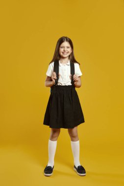 Neşeli bir çocuk okul üniforması içinde duruyor, sarı bir arka planda sırt çantasıyla gülümsüyor. Eğitici pazarlama materyalleri ve sosyal medya içeriği için ideal.