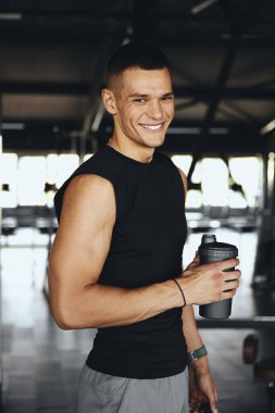 Spor salonunda elinde protein karıştırıcı tutarken kameraya gülümseyen kendine güvenen genç bir adam, egzersiz sonrası. Sağlıklı yaşam tarzı, spor programı ve aktif erkek model kavramı.