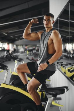Geniş modern spor salonunda bisiklet makinesinde otururken pazılarını gösteren ciddi bir adamın dikey fotoğrafı..