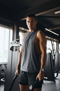 İyi donanımlı bir spor salonunda savaş halatları tutan atletik bir adamın dikey fotoğrafı, dayanıklılık ve güç gösterisi. Sporcu erkek fitness merkezinde halatla poz veriyor.