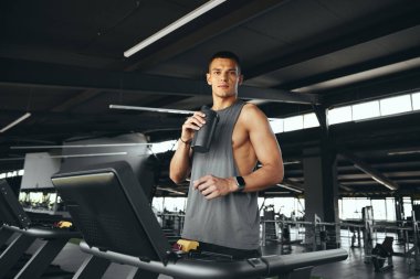 Aktif, gri tişörtlü, şişeden su içen koşu bandında duran çekici bir genç. Spor seansından sonra susuz kalmayan genç bir adam. Sağlıklı yaşam tarzı, spor salonunda erkek eğitimi.. 