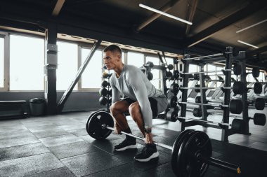 Ağırlık rafları ve ekipmanlarla çevrili modern bir spor salonunda halterle kaldırma yapan odaklanmış bir sporcu. Güç eğitimi, disiplin ve iş başındaki yaşam tarzı.