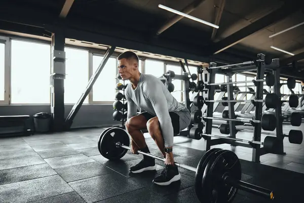 Ağırlık rafları ve ekipmanlarla çevrili modern bir spor salonunda halterle kaldırma yapan odaklanmış bir sporcu. Güç eğitimi, disiplin ve iş başındaki yaşam tarzı.