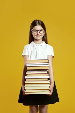 Gözlük takan neşeli bir çocuk öğrenme hevesini somutlaştıran parlak sarı bir arkaplan önünde kendinden emin bir şekilde uzun bir kitap yığını tutuyor. Eğitici içerik promosyonları için ideal.