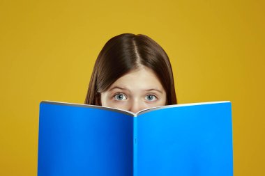 Meraklı bir çocuk parlak mavi bir kitaba bakar, geniş gözleri odaklanmış ve meraklıdır. Parlak sarı arka plan merak duygusunu arttırır. Eğitim içeriği ve okul promosyonları için ideal.