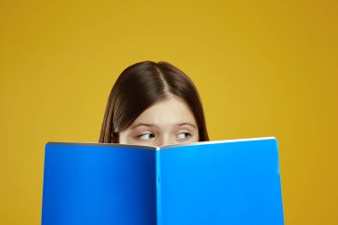 Meraklı bir çocuk, parlak mavi bir kitaba bakıyor. Parlak sarı bir arkaplana bakıyor. Merakını ve dikkatini gösteriyor. Eğitim içeriği ve pazarlama materyalleri için ideal.