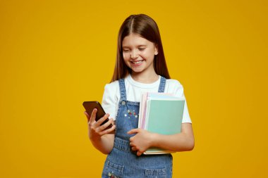 Kot elbiseli, akıllı telefon kullanan, kitapları tutan, stüdyo ortamında gülümseyen mutlu çocuk. Eğitimsel promosyonlar ve teknolojik pazarlama görselleri için ideal.