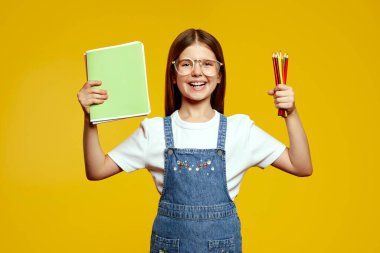 Renkli kalemler ve defterler tutan şık kot tulumlu ve moda gözlüklü zeki ve yakışıklı liseli kız. Neşeli gülüşü ve parlak geçmişi öğrenme hevesine ışık saçıyor.