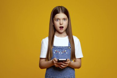 Kot pantolon giyen meraklı bir çocuk, sarı arka planda izole edilmiş bir akıllı telefonu tutarken şaşkınlığını dile getiriyor. Reklam ve teknik promosyonlar için ideal..