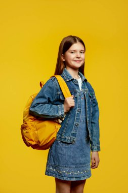 Gülümseyen genç çocuk, elinde sarı bir sırt çantası, şık bir kot giysisi, neşe ve hazırlığı simgeliyor. Okul promosyonları veya gençlik moda reklamları için ideal.