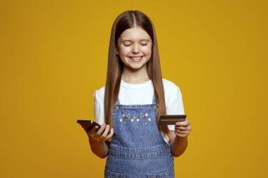 Gülümseyen çocuk neşeli bir şekilde bir telefon ve kredi kartı tutuyor, heyecan ve masumiyet saçıyor. Dijital teknoloji ve bankacılık hizmetlerinde terfi için ideal.