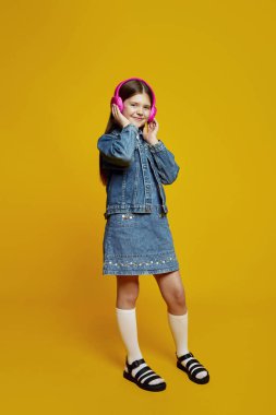 Kot elbiseli ve ceketli gülümseyen genç, pembe kulaklıklardan canlı sarı bir arka plana rağmen hoş bir şekilde müzik dinliyor. Çocuklar için ideal moda reklamcılığı veya yaşam tarzı pazarlaması