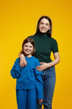 Dişleriyle gülümseyen ve kucaklaşan sevimli kız kardeşler aile portresi için poz verirken sarı arka planda duruyorlar.