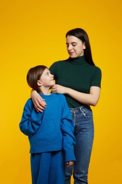 Gülümseyen güzel bir kadın ve küçük kız kardeş birbirlerine bakıyorlar, sarı arka plan stüdyosunda soyutlanmışlar.